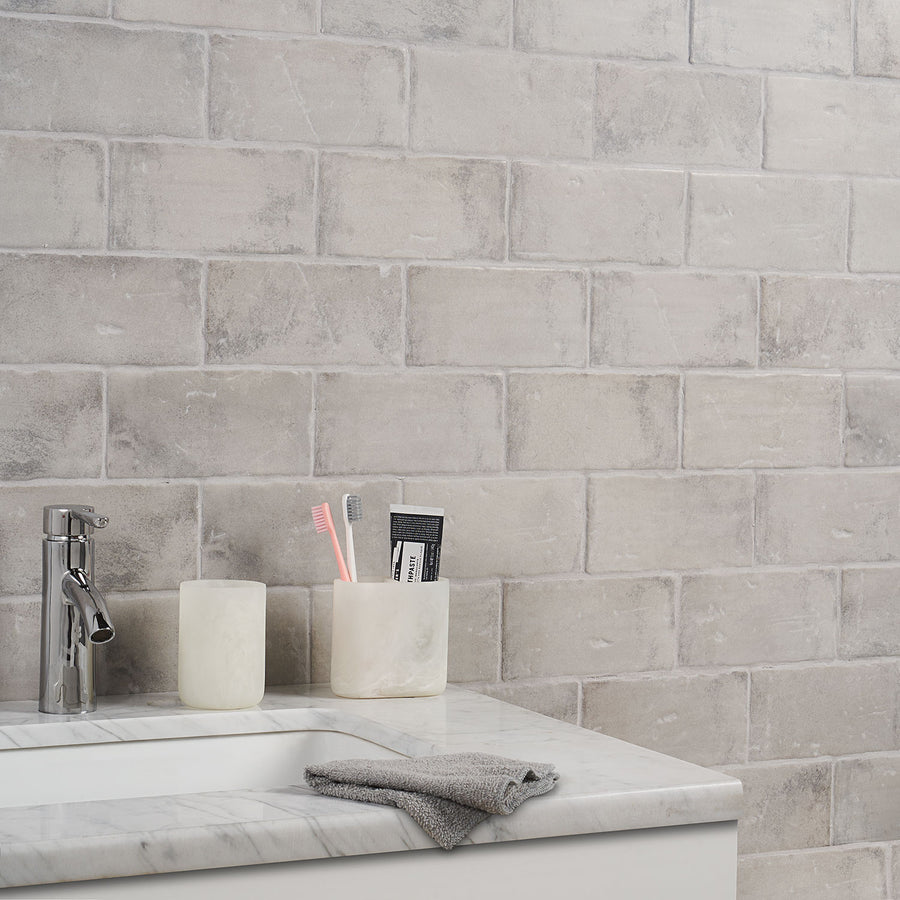 A marble bathroom counter with a silver faucet, a gray washcloth, and two marble holders—one with a pink toothbrush, a gray toothbrush, and toothpaste, and the other empty—in front of a light gray tiled wall.