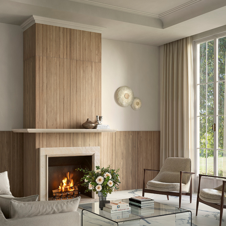 A modern living room with a lit fireplace, light wood paneling, Corsica Fluted 24x48 Ceramic Tile accents, neutral armchairs, a glass coffee table with books and flowers, and large windows with sheer curtains letting in natural light.