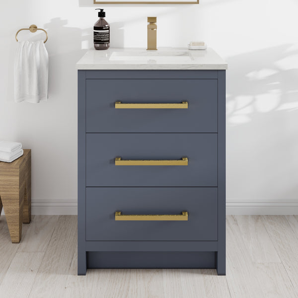 A dark blue Curio 24″ Vanity featuring three gold handles, a Carrara quartz top, a sink, a soap dispenser, a towel ring with a white towel, and a wooden stool with folded towels sits elegantly on a light wood floor—perfect for creating a stylish modern bathroom vanity setup.