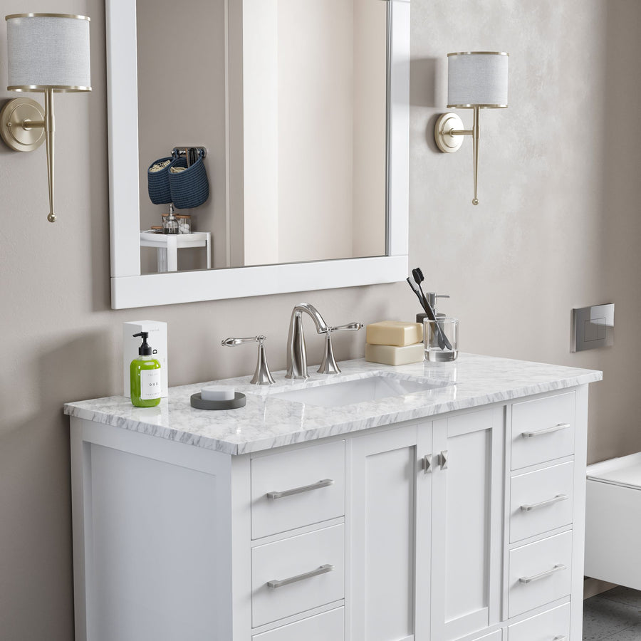 A modern bathroom vanity with a white countertop, sink, faucet, various toiletries, and a soap dispenser. Above is a large mirror, with wall sconces on either side, and a towel or basket reflected in the mirror.