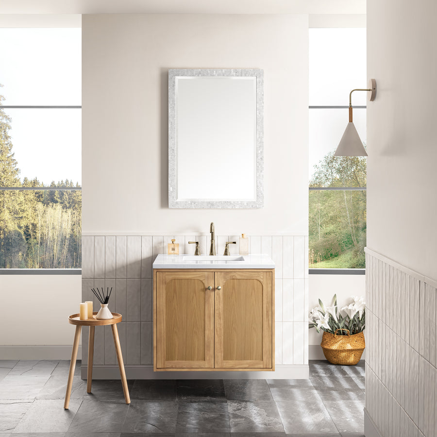A modern bathroom with the Laurent 30" Vanity in natural oak, white sink, large mirror, wall light, a round table with decor, and a basket of white flowers by the window showcasing scenic outdoor views.