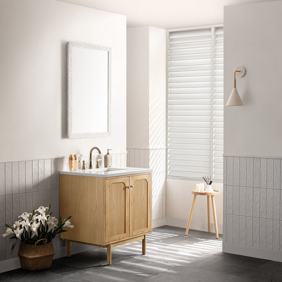 Minimalist bathroom with a wooden vanity, marble countertop, and rectangular mirror. A basket of white flowers sits on the floor. Sunlight streams through white blinds, illuminating a stool and modern wall sconce.