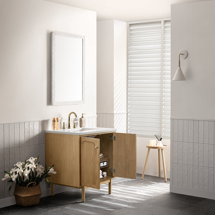 A modern bathroom with a wooden vanity, white countertop, and open cabinet doors. A basket of white lilies sits on the floor, next to a window with white blinds and a wooden stool. A wall sconce and mirror are above the sink.
