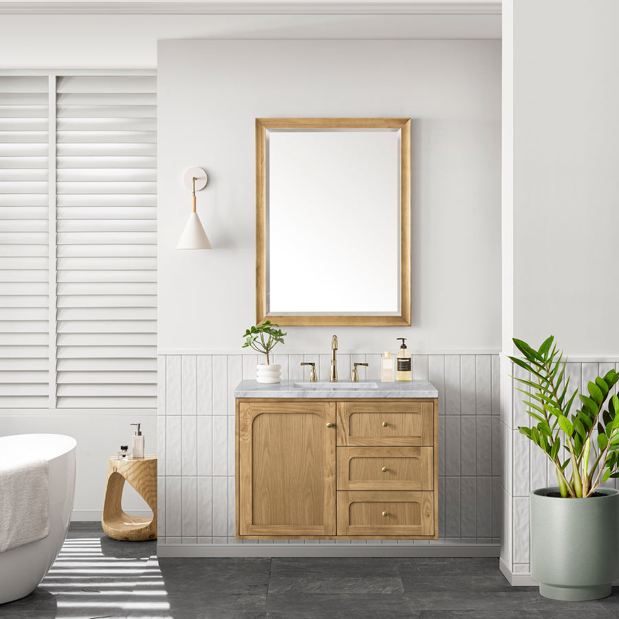 A modern bathroom showcases the Laurent 36" Vanity in natural oak, a rectangular mirror, wall-mounted light, lush plants, white walls, gray tiles, and a freestanding tub beside shuttered windows.