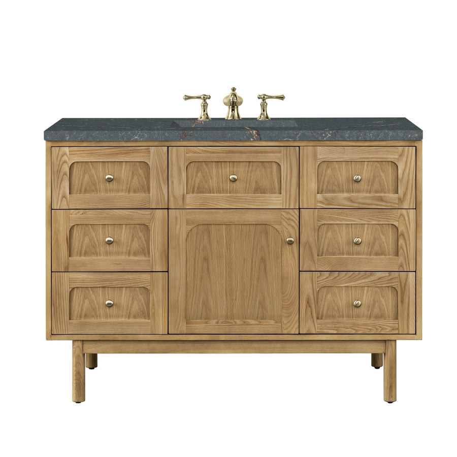 A wooden bathroom vanity with a black countertop, brass faucet fixtures, and multiple drawers and a central cabinet for storage. The design is modern with a natural wood finish.