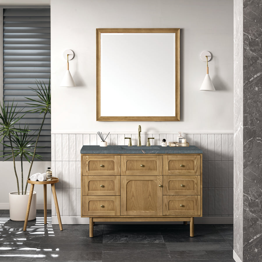 A modern bathroom features the Laurent 48" Vanity, natural oak with a gray top, paired with a large mirror, wall sconces, and a small stool holding a plant and toiletries. Tiled walls and a dark stone floor finish the space.
