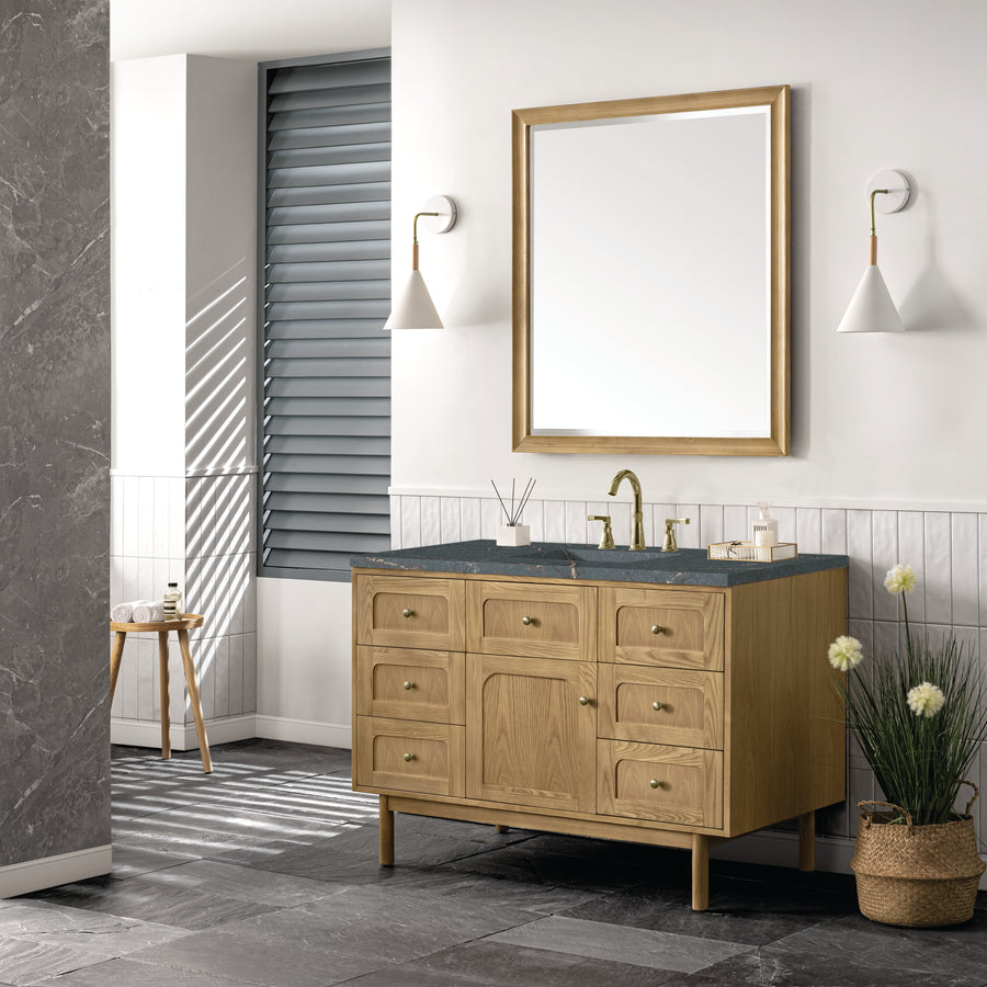 Modern bathroom with a wooden vanity, black countertop, gold faucet, large mirror, two white sconces, potted plant, and a stool by a window with slatted blinds. Sunlight creates shadows on the floor and wall.