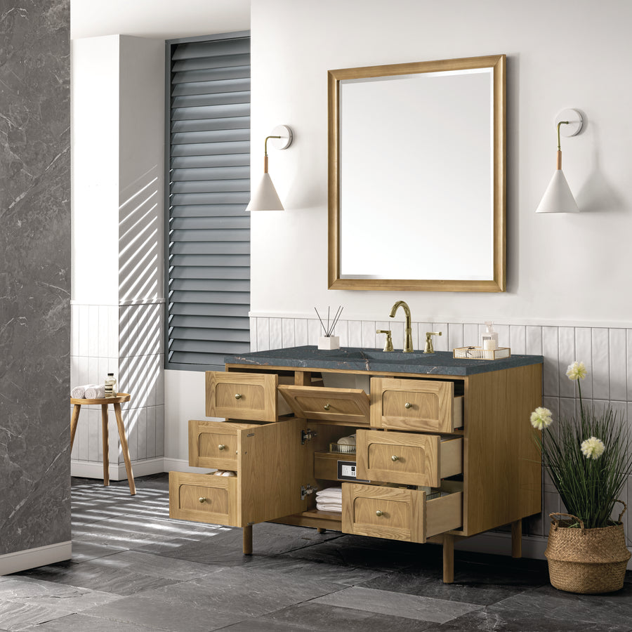 Modern bathroom with a wooden vanity featuring open drawers and doors, a black countertop, brass faucet, large mirror, wall sconces, a stool, plant, and gray tiled floor. Light streams in through window blinds.