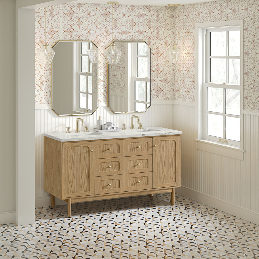 A bathroom with a wood vanity, double sinks, gold fixtures, two geometric mirrors, patterned tile floor, wallpaper with a pink floral design, and a large window letting in natural light.