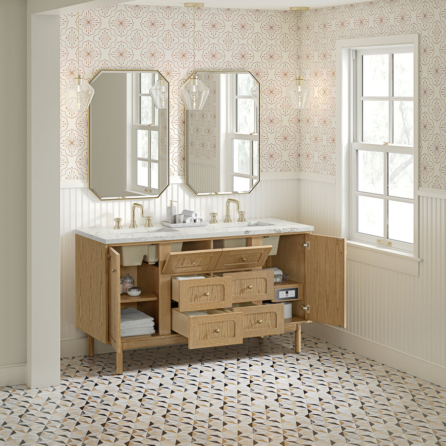 A bright bathroom with two mirrors, double sink, light wood vanity with drawers and cabinets open, gold fixtures, geometric patterned floor, and wallpaper with a pink and gold design. A window lets in natural light.