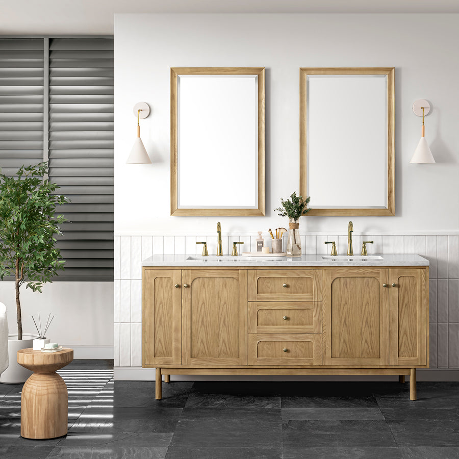 Modern bathroom with the Laurent 72" Vanity Double Bowl in light oak, two rectangular mirrors, brass fixtures, wall sconces, a small counter plant, and a potted plant beside a wooden stool on dark tile floors.