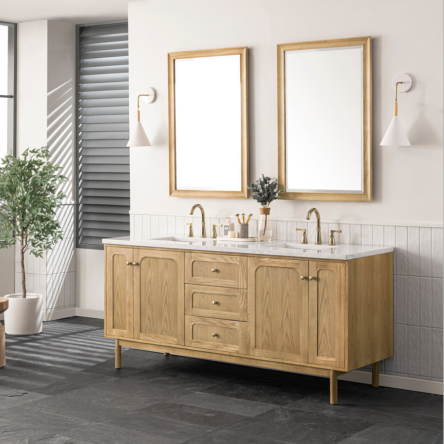 A modern bathroom with a double-sink wooden vanity, two rectangular mirrors, gold faucets, white wall sconces, a potted plant, and gray tile flooring. The room has white and light gray walls.