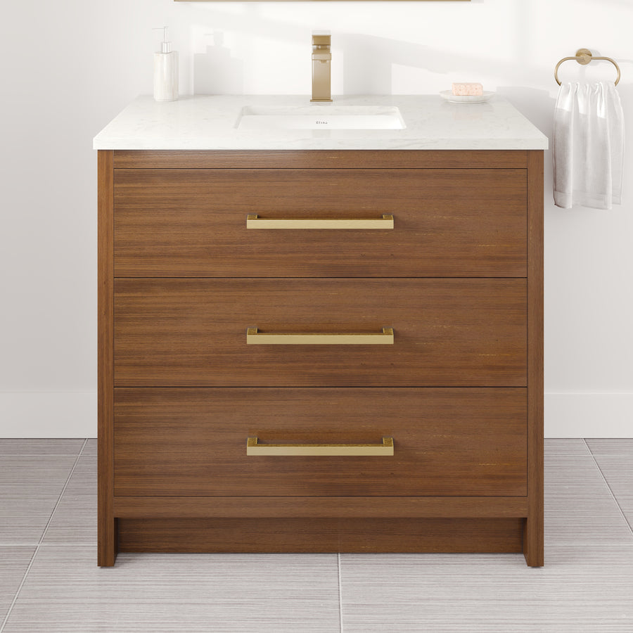 A Curio 36″ Vanity in luxurious walnut features three drawers with elegant gold handles, topped with a pristine Carrara quartz surface that includes a built-in sink. The bathroom vanity is complete with a soap dispenser, soap, and a towel ring holding a white towel on the right.