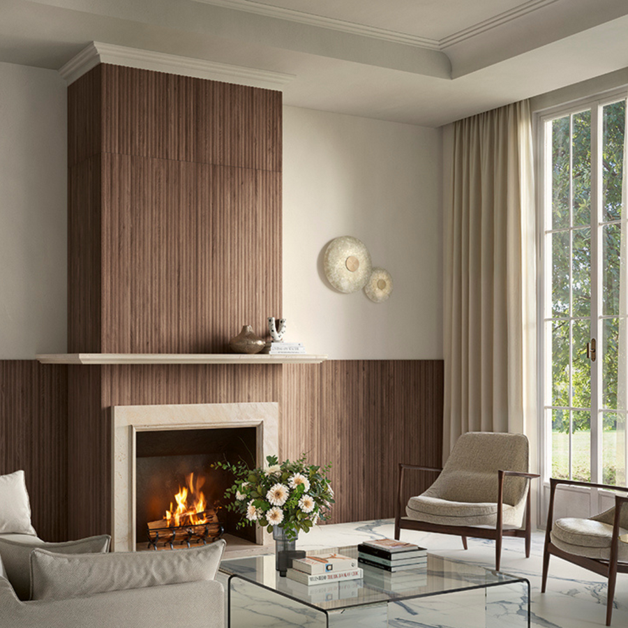 A modern living room with a lit fireplace, a wood-panel accent wall featuring Corsica Fluted 24x48 Ceramic Tile, a marble coffee table with flowers, two armchairs, and large windows with cream curtains overlooking greenery.