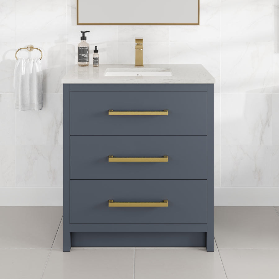 The Curio 30″ Vanity showcases a stunning dark blue bathroom vanity with three gold-handled drawers, enhanced by a pristine Carrara quartz top and paired with a rectangular sink. The refined gold faucet, matching soap dispenser, and towel ring holding a white towel beautifully complement the marble-tile walls and floor, creating a fresh, contemporary ambiance.