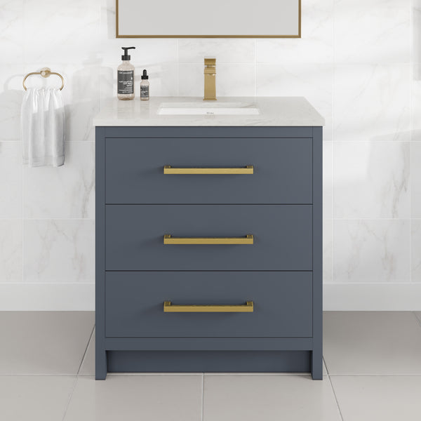 The Curio 30″ Vanity showcases a stunning dark blue bathroom vanity with three gold-handled drawers, enhanced by a pristine Carrara quartz top and paired with a rectangular sink. The refined gold faucet, matching soap dispenser, and towel ring holding a white towel beautifully complement the marble-tile walls and floor, creating a fresh, contemporary ambiance.