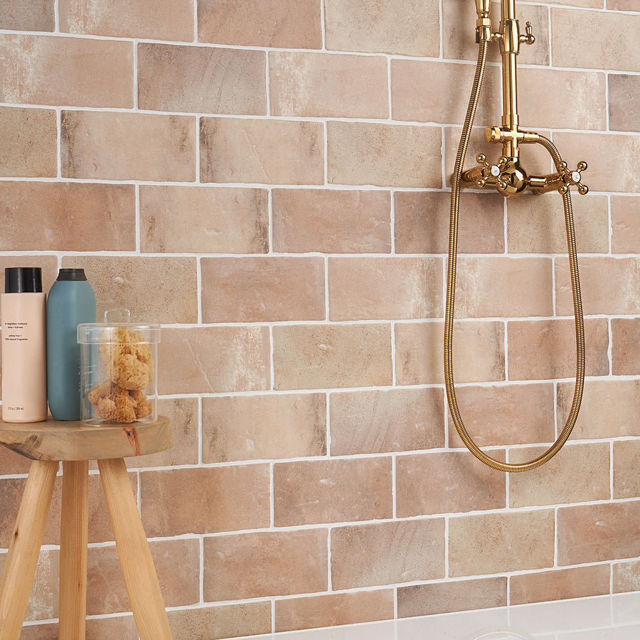 A shower area with tan and beige brick-style tiles, a vintage brass shower fixture, and a wooden stool holding a blue bottle, a beige bottle, and a clear jar with natural sponges.