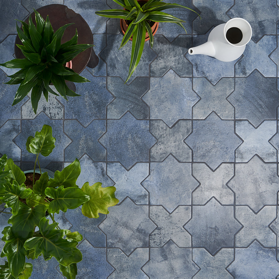 Three green potted plants and a white watering can sit on a patterned blue and gray tile floor viewed from above. The tiles form a repeating geometric star-shaped design.