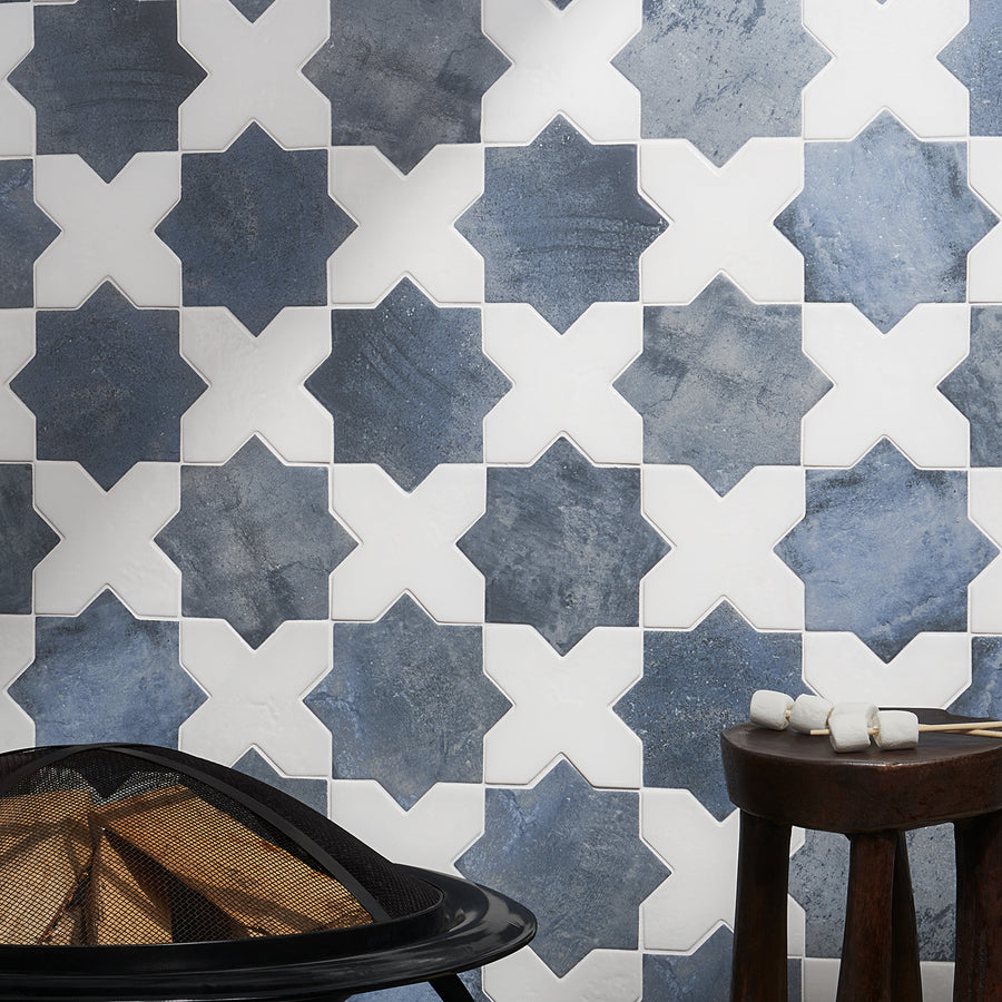 A wall with a geometric tile pattern featuring alternating blue and white star-shaped tiles. A wooden stool with marshmallows on sticks and a black fire pit are in the foreground.