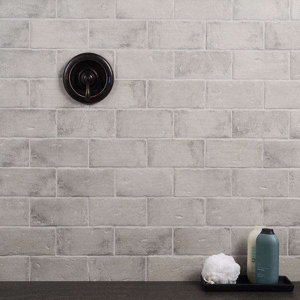 A shower wall with light gray, rectangular tiles, a round black metal shower control handle, and a black tray holding a white bath sponge and bottles, set on a dark countertop.