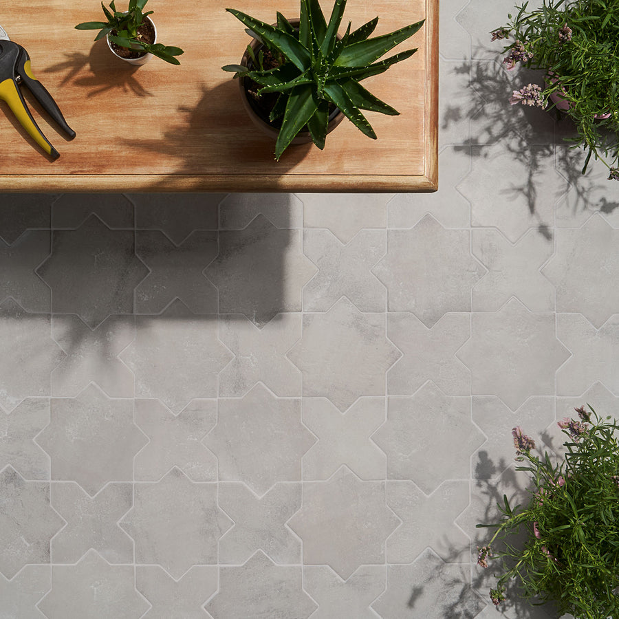 A wooden table with potted plants and pruning shears sits on a light gray, intricately patterned tile floor. More potted plants are visible in the corners of the image.