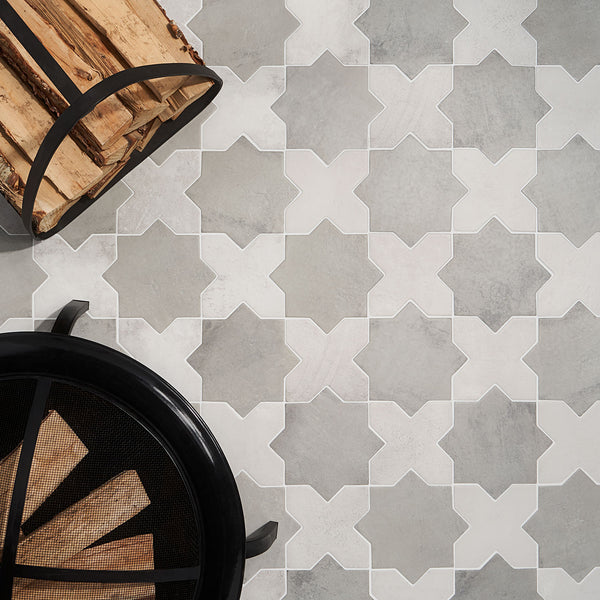 Geometric gray and white tile floor with star-shaped patterns, next to a black metal firewood holder filled with logs and the edge of a black metal fireplace screen.