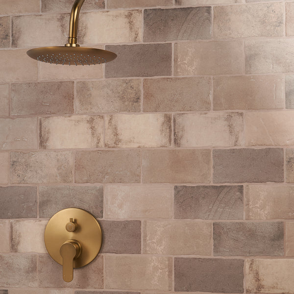 A shower with a round brass showerhead and matching handle is mounted on a wall covered in beige and brown rectangular tiles arranged in a staggered pattern.