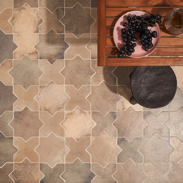 A table with a pink plate of dark grapes, a glass, and a wooden stool sits on a floor with beige and brown star-shaped patterned tiles.