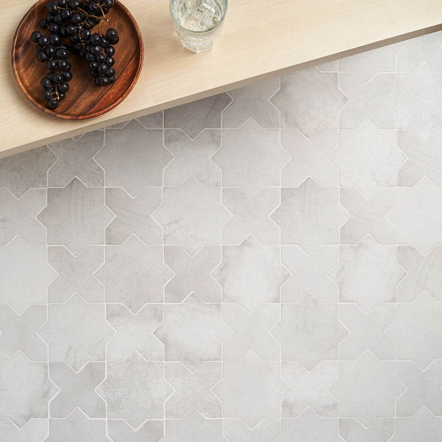 A light gray, geometric tile floor with a star-shaped pattern is shown. On a light wooden surface above, there is a glass of water and a wooden plate with a bunch of dark grapes.