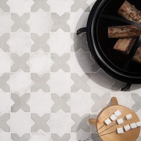 Overhead view of patterned tile flooring with geometric shapes, a round fire pit holding firewood, and a small round wooden table with skewers of marshmallows ready for roasting.