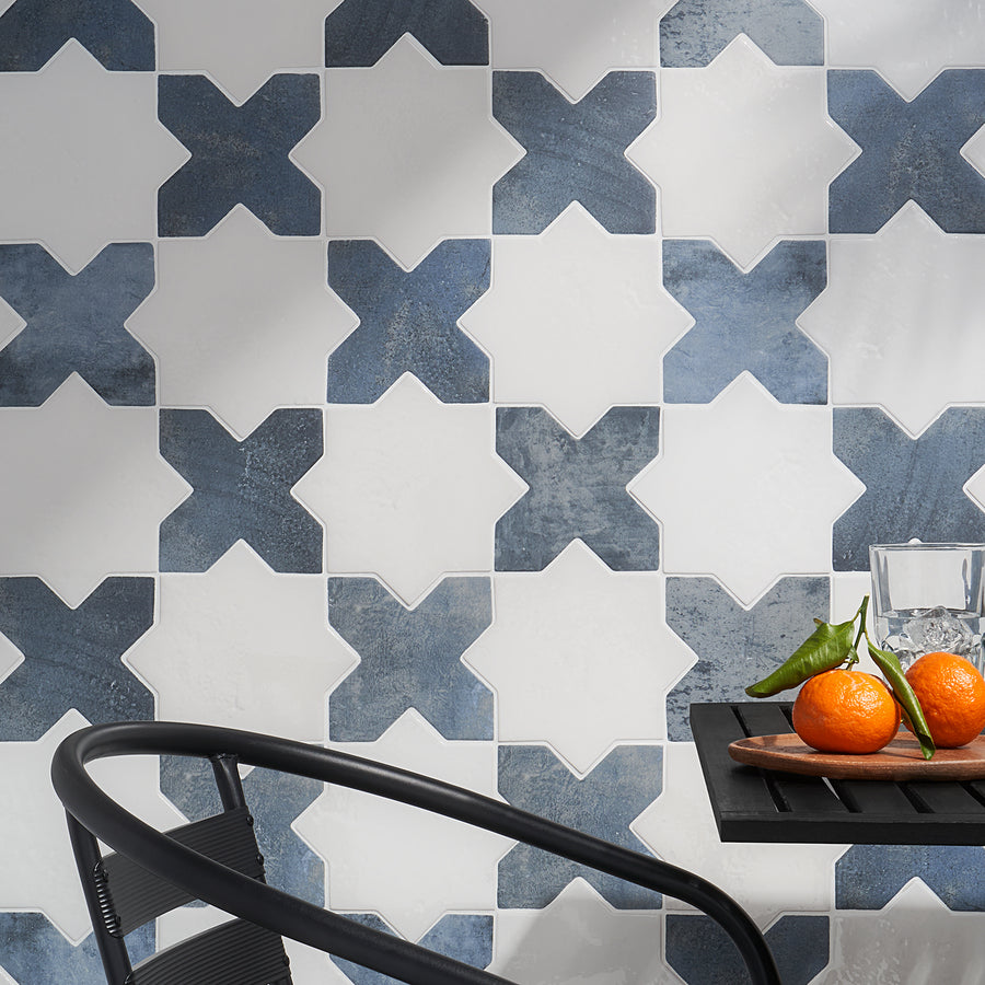 A wall with a repeating tile pattern featuring white and blue star shapes. In the foreground, a black chair and a black table hold two tangerines, a green chili, and a glass of water.