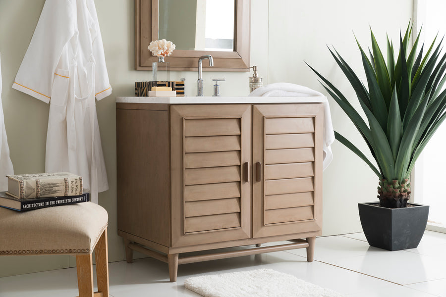 A modern bathroom with a wooden vanity, a rectangular mirror, a potted plant, a white towel, and a robe hanging on the wall. A cushioned stool with books is nearby, and the floor is white tile with a small rug.