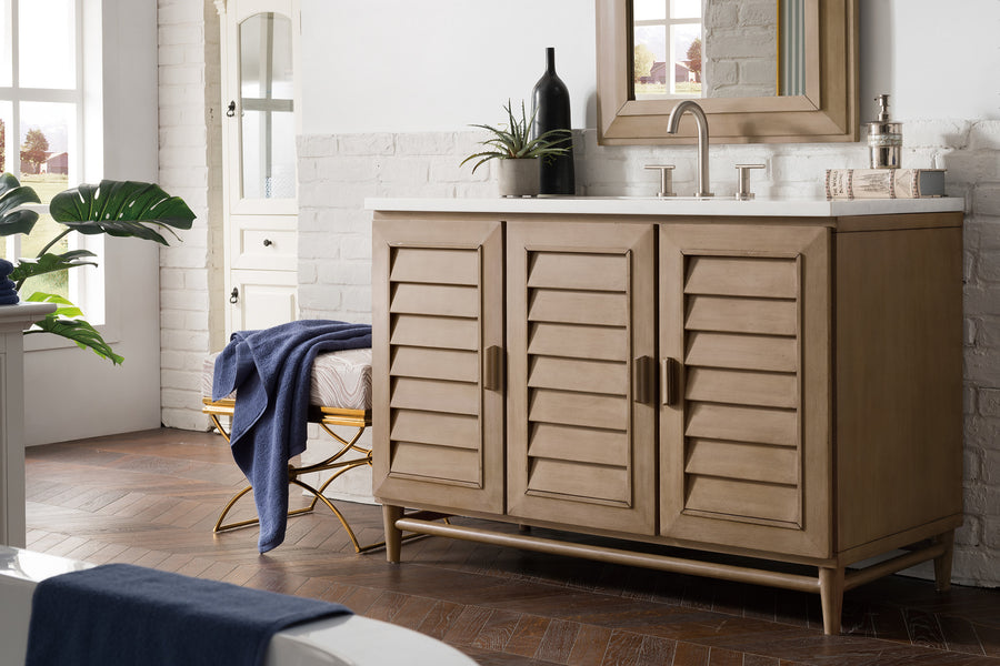 A modern bathroom features a wooden vanity with shutter-style doors, a countertop sink, a mirror, and various decorative items. A blue towel is draped over a bench nearby, and plants add greenery to the space.