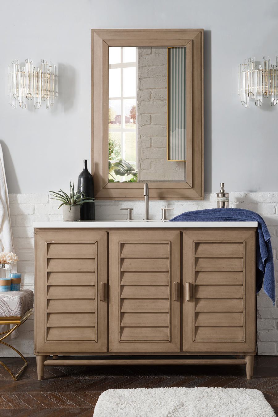 The Portland 48" Vanity boasts a modern wood finish, louvered doors for spacious storage, a sleek rectangular mirror above, silver faucet, side sconces, stylish accents, and a neatly hung blue towel on the right.