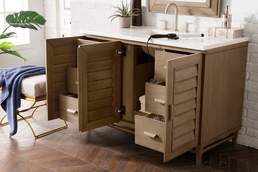 A wooden bathroom vanity with louvered cabinet doors and open drawers shows towels and toiletries inside. The sink countertop has a faucet, a plant, books, and a curling iron.