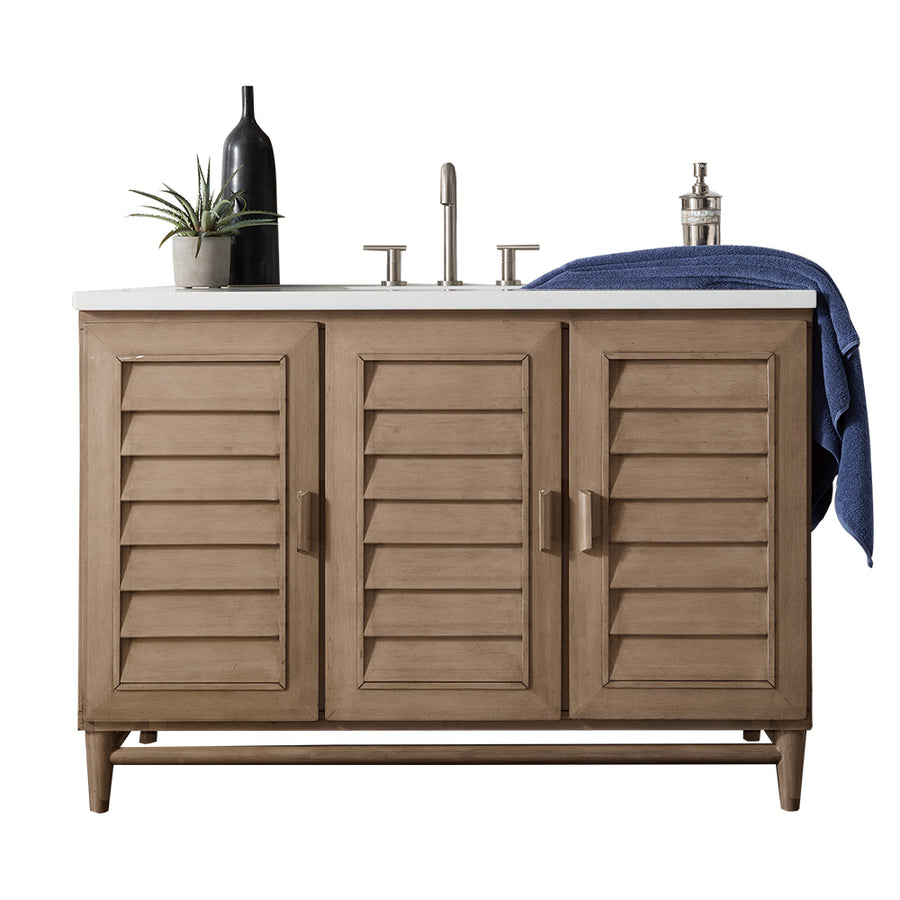 A wooden bathroom vanity with three louvered cabinet doors, a white countertop, sink with faucet, a potted plant, a dark bottle, a soap dispenser, and a blue towel draped on the right side.