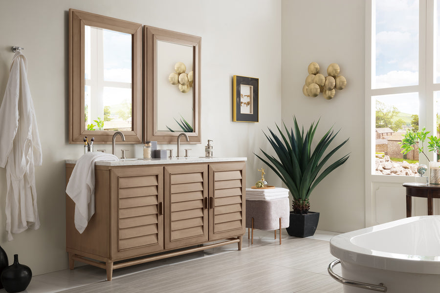 Modern bathroom with a double-sink wooden vanity, two mirrors, gold wall sconces, a large potted plant, robe on a hook, a framed picture, and a freestanding bathtub by tall windows letting in natural light.