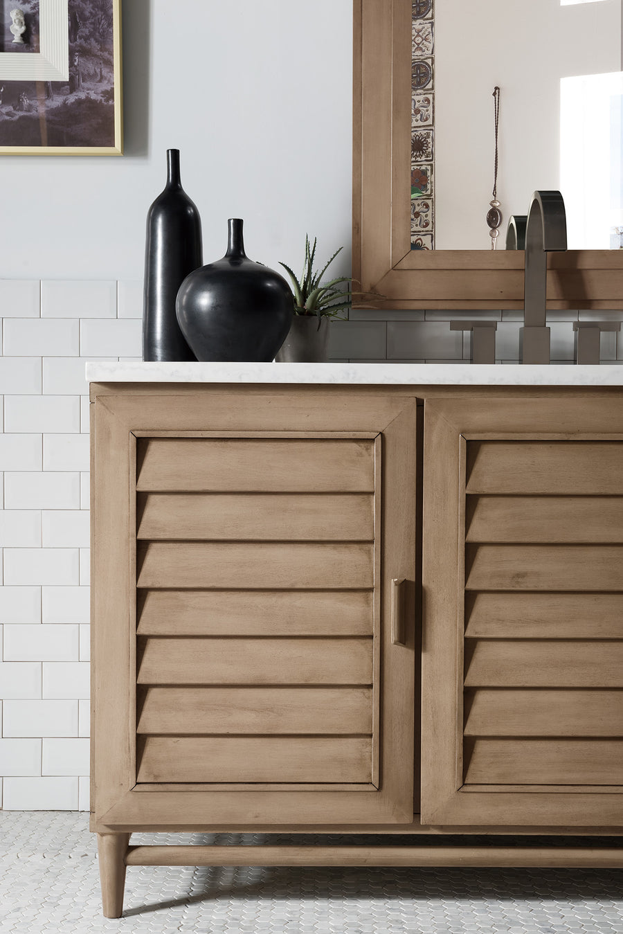 Light wood bathroom vanity with louvered doors, white countertop, two black decorative vases, and a small plant. Rectangular mirror and white tiled wall in the background.