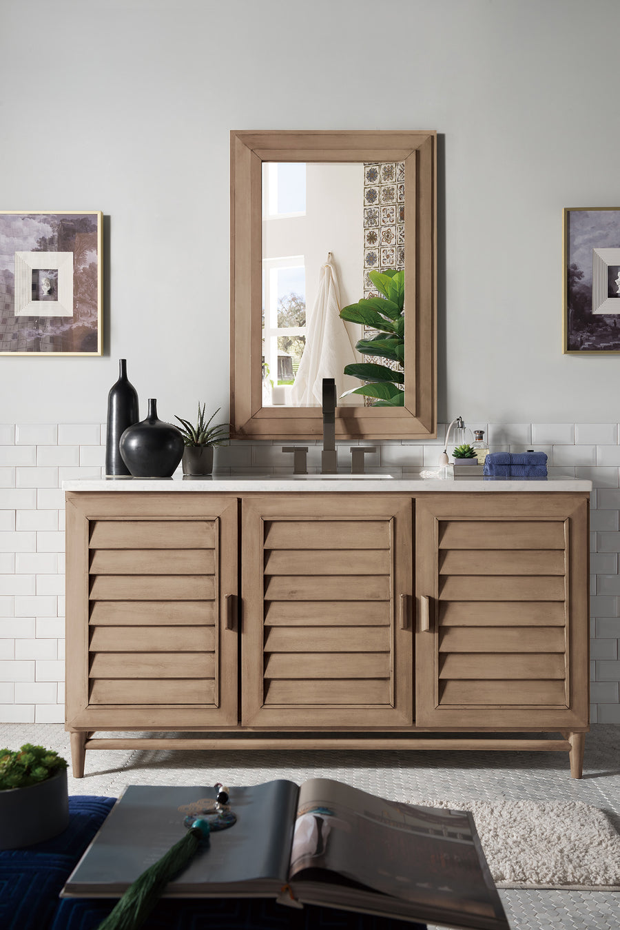 The Portland 60" Vanity features louvered cabinet doors for stylish bathroom storage, topped with a white counter and minimalist decor beneath a coordinating mirror. White subway tiles and framed art complement the light-toned wall.