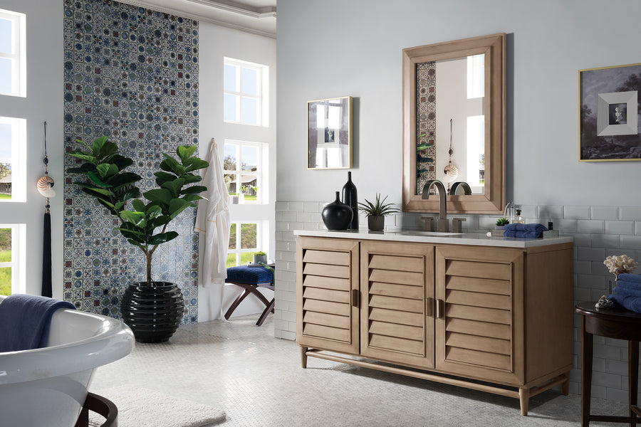 Bright bathroom with a wooden vanity, a large potted plant, a framed mirror, blue mosaic accent wall, white clawfoot tub, and multiple windows letting in natural light. Navy towels and small decor items are placed on the vanity.