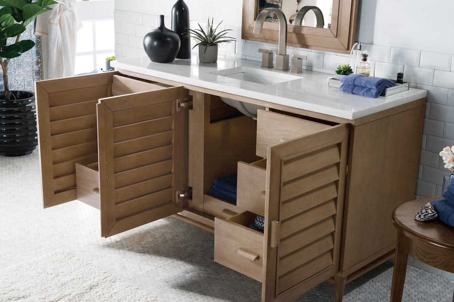 A bathroom vanity with light wood cabinets and drawers open, revealing storage space. The countertop holds a sink, faucet, two black vases, a soap dispenser, a plant, and folded blue towels.