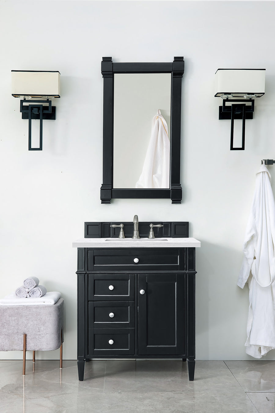 A modern bathroom features the Brittany 30" Vanity, a matching mirror, chrome faucet, wall lights, a towel rack with a white robe, and neatly folded towels on a gray bench.