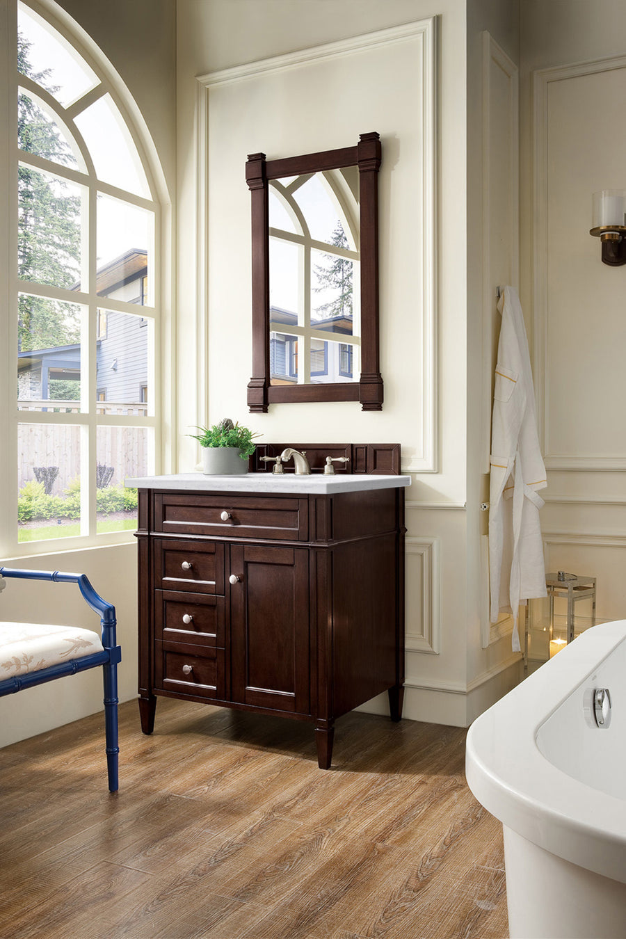 A bright bathroom showcases the Brittany 30" Vanity, with a large arched window, matching mirror, towel on a hook, wood floors, and part of a white bathtub in the foreground.
