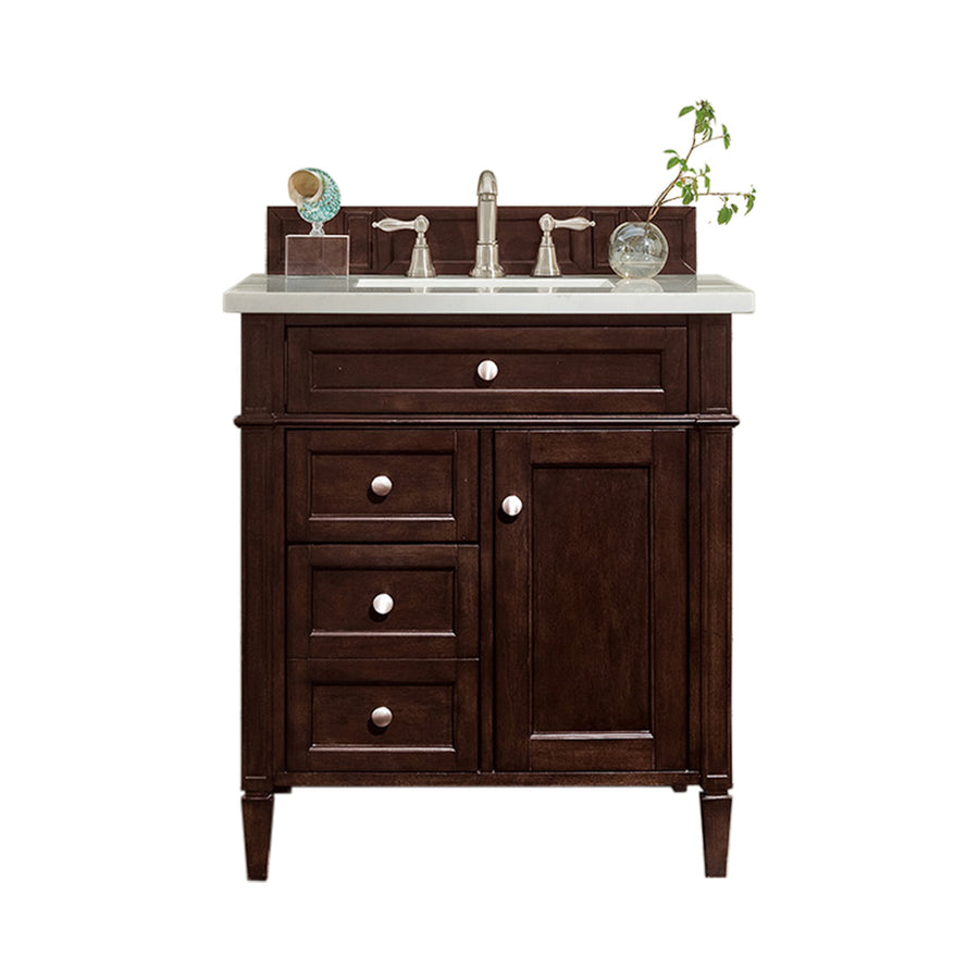 A dark wood bathroom vanity with a white countertop, silver faucet, three small drawers, one cabinet door, and a small plant and decorative items on the counter.
