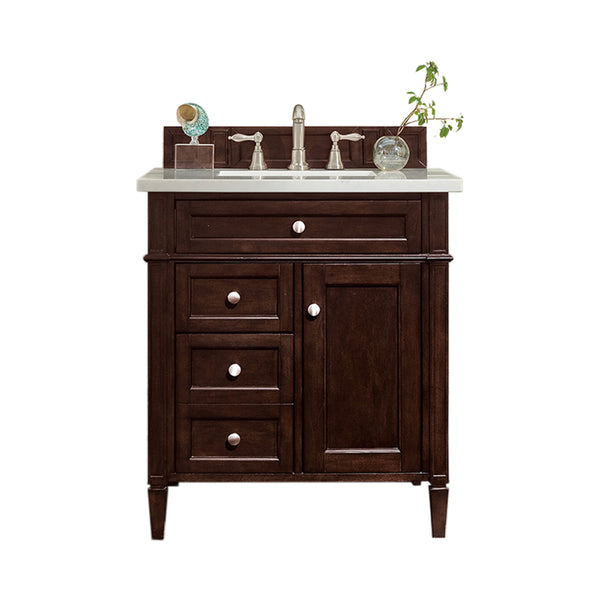 A dark wood bathroom vanity with a white countertop, silver faucet, three small drawers, one cabinet door, and a small plant and decorative items on the counter.
