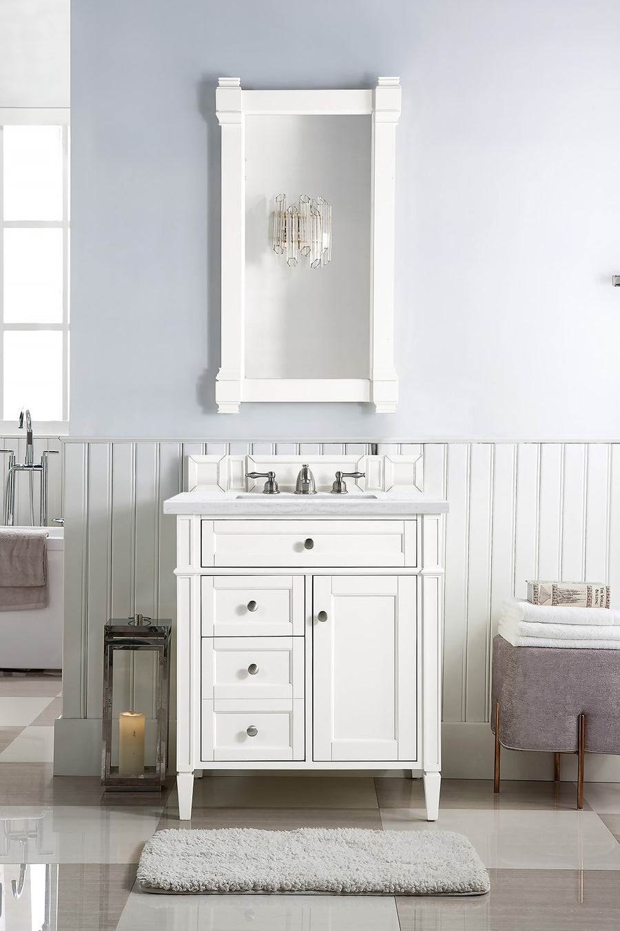 A modern white bathroom features the Brittany 30" Vanity with drawers, a rectangular mirror, wall sconce, plush rug, candle lantern on the floor, and a cushioned bench against soft blue and white paneled walls.
