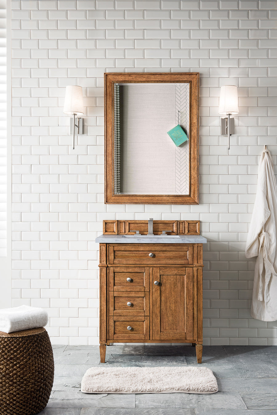 The Brittany 30" Vanity with a gray top sits beneath a framed mirror on white tile. Two wall sconces, a robe, woven hamper, and white rug complete the clean, modern look.