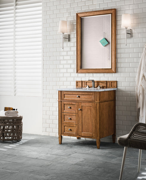 A bathroom with white subway tile walls, a wooden vanity with a black countertop, a large framed mirror, wall sconces, a wicker laundry basket, a chair, and sunlight streaming through white shutters.
