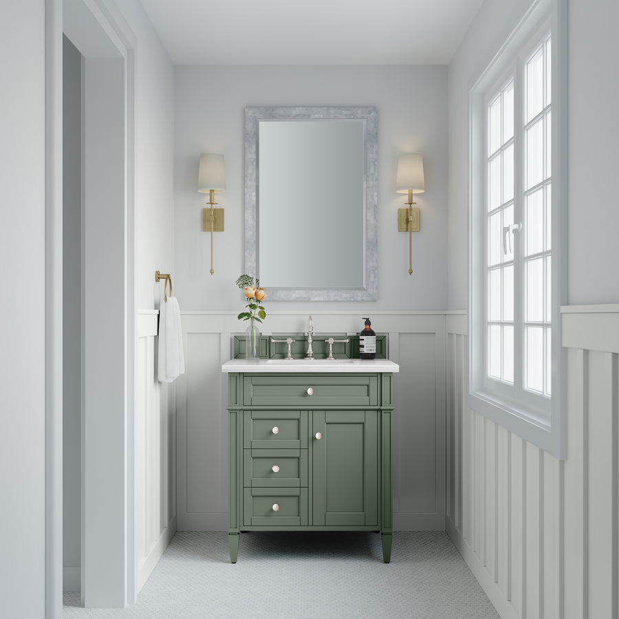 A bathroom features a Brittany 30" Vanity (green) with a white countertop, gold fixtures, large mirror, wall sconces, towel ring with white towel, natural light from a window, and elegant white wainscoted walls.
