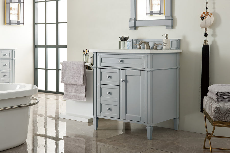 A modern bathroom with light gray vanity, white countertop, silver faucet, a mirror above, towel rack with a light purple towel, decorative items, and marble flooring. A bathtub and stacked towels are also visible.